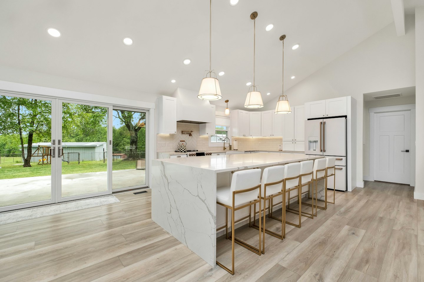 Modern white kitchen with marble island and bar stools opens to green backyard.